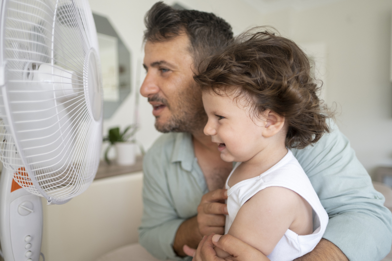 Father and toddler sitting in front of a fan in their living room on a hot day.