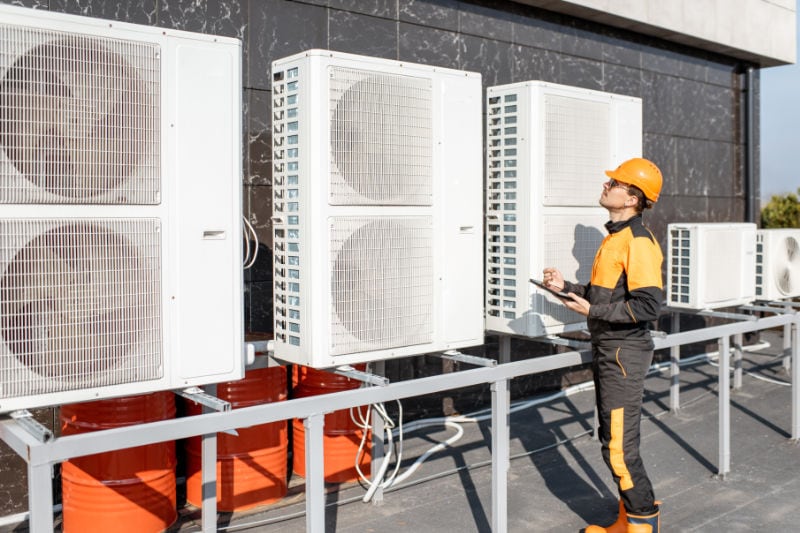 AC tech wearing yellow and black gear holding a clipboard and pen analyzing outdoor white commercial hvac equipment on top of the roof of a commercial building on a sunny day.