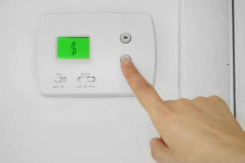 Revolutionize Your Home Comfort With a Smart Thermostat. Person adjusting a wall thermostat with dollar sign symbol on the display.